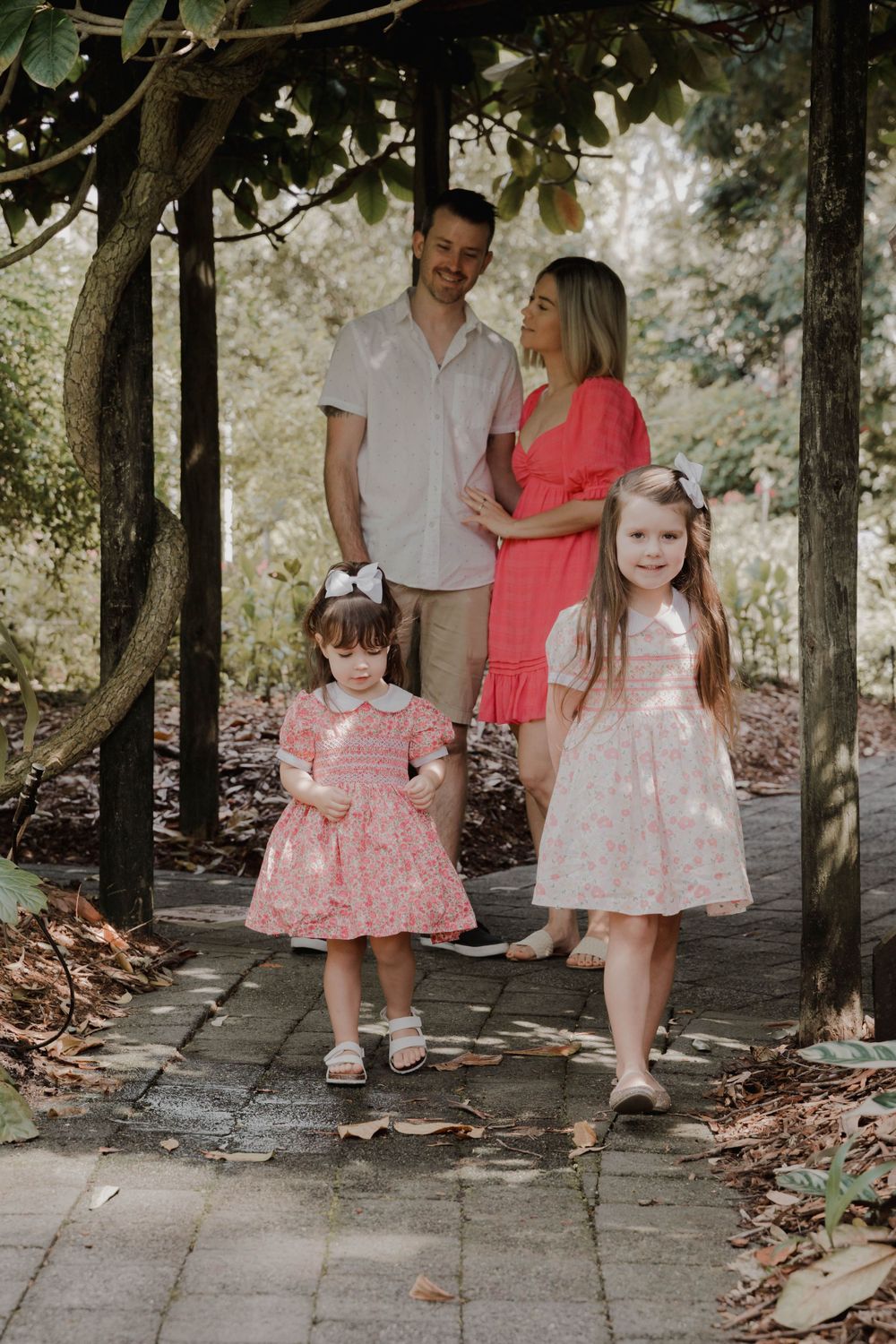 A family of four poses together on a garden path wearing coordinating pink and beige outfits.
