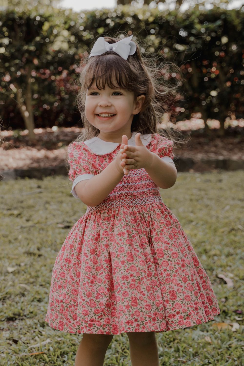 A little girl in a floral dress and bow twirls happily in a garden setting.