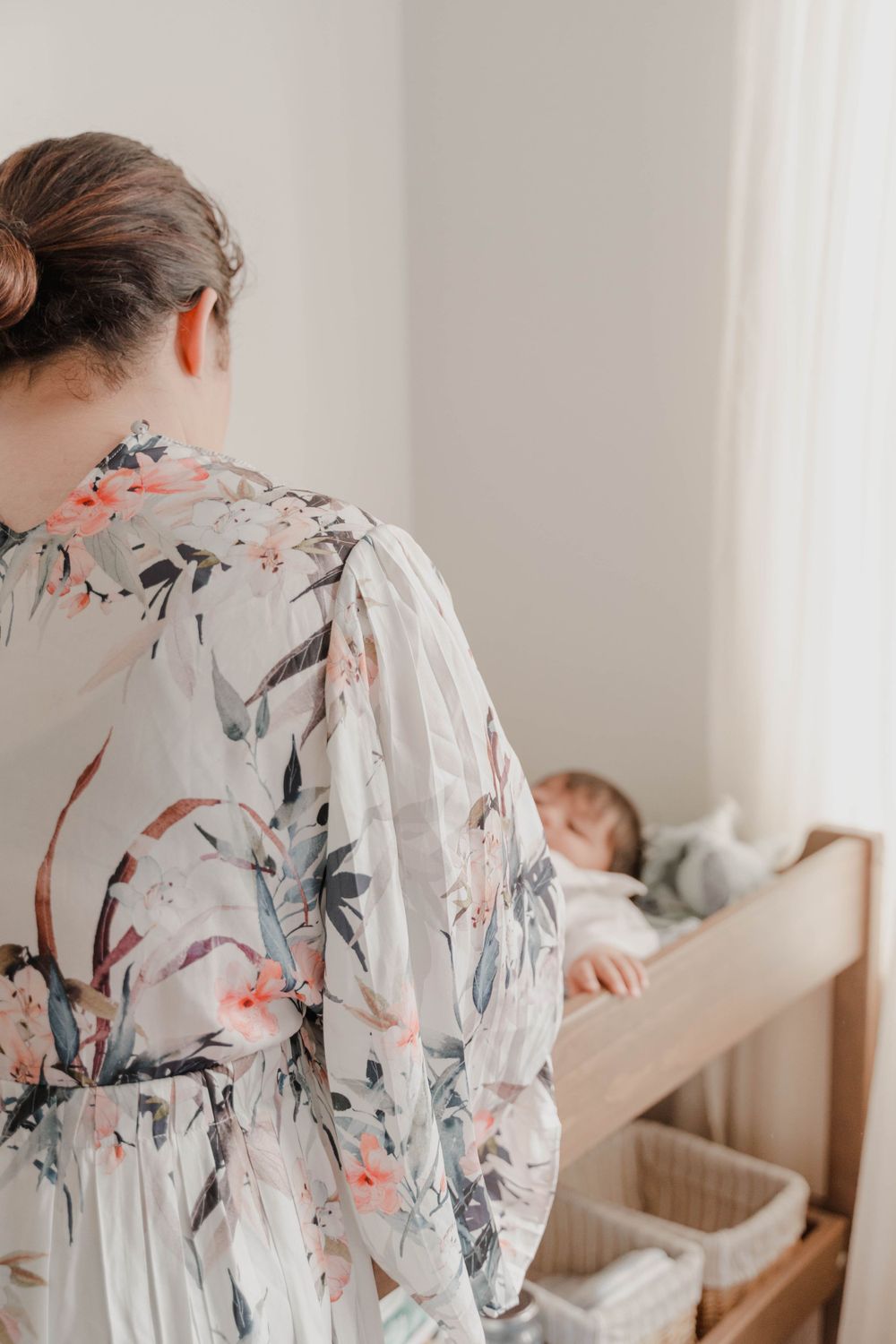 A person in a floral robe stands near a child sleeping in bed.