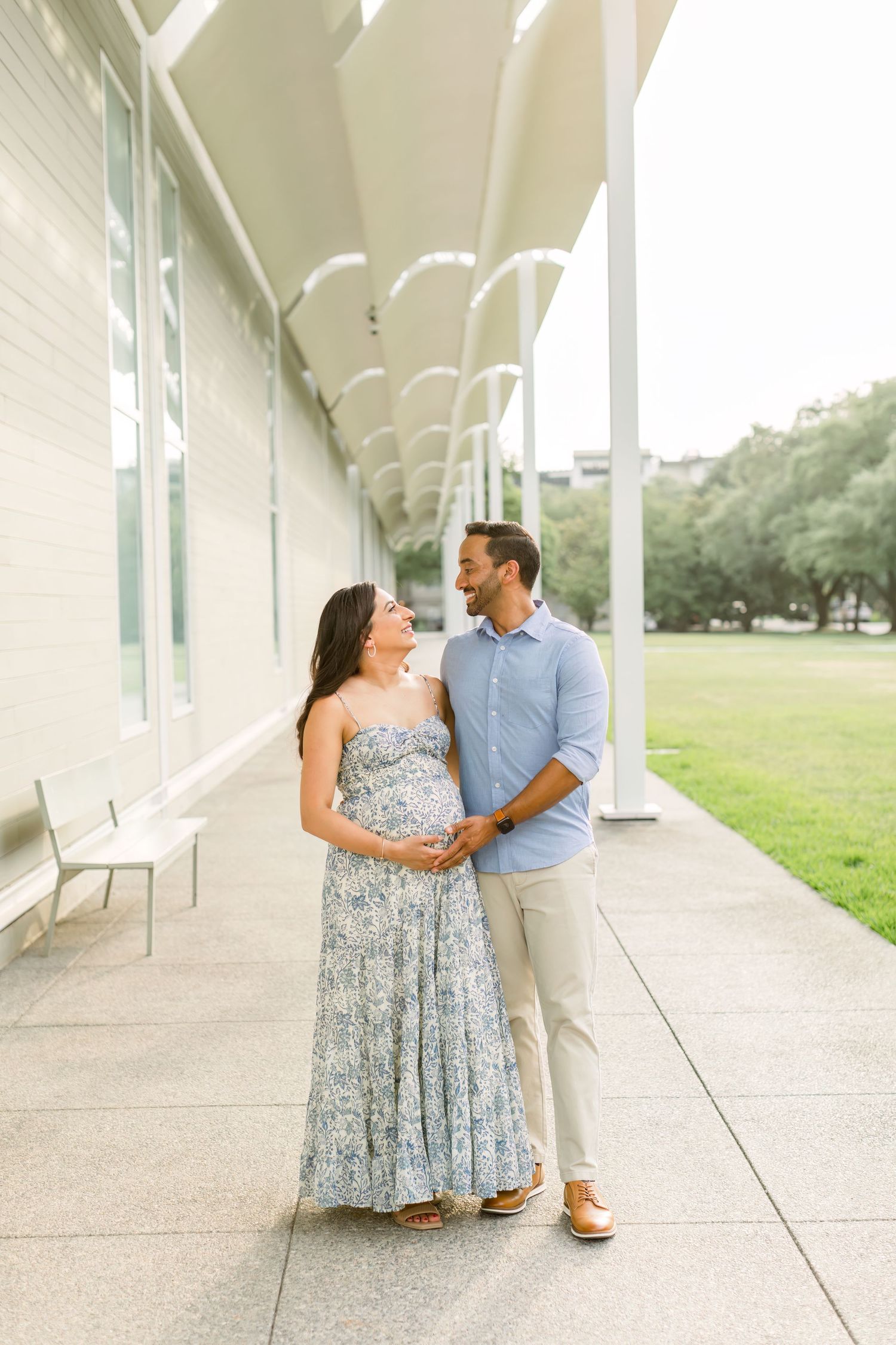 Expectant couple sharing a tender moment under a white columned portico in formal attire.