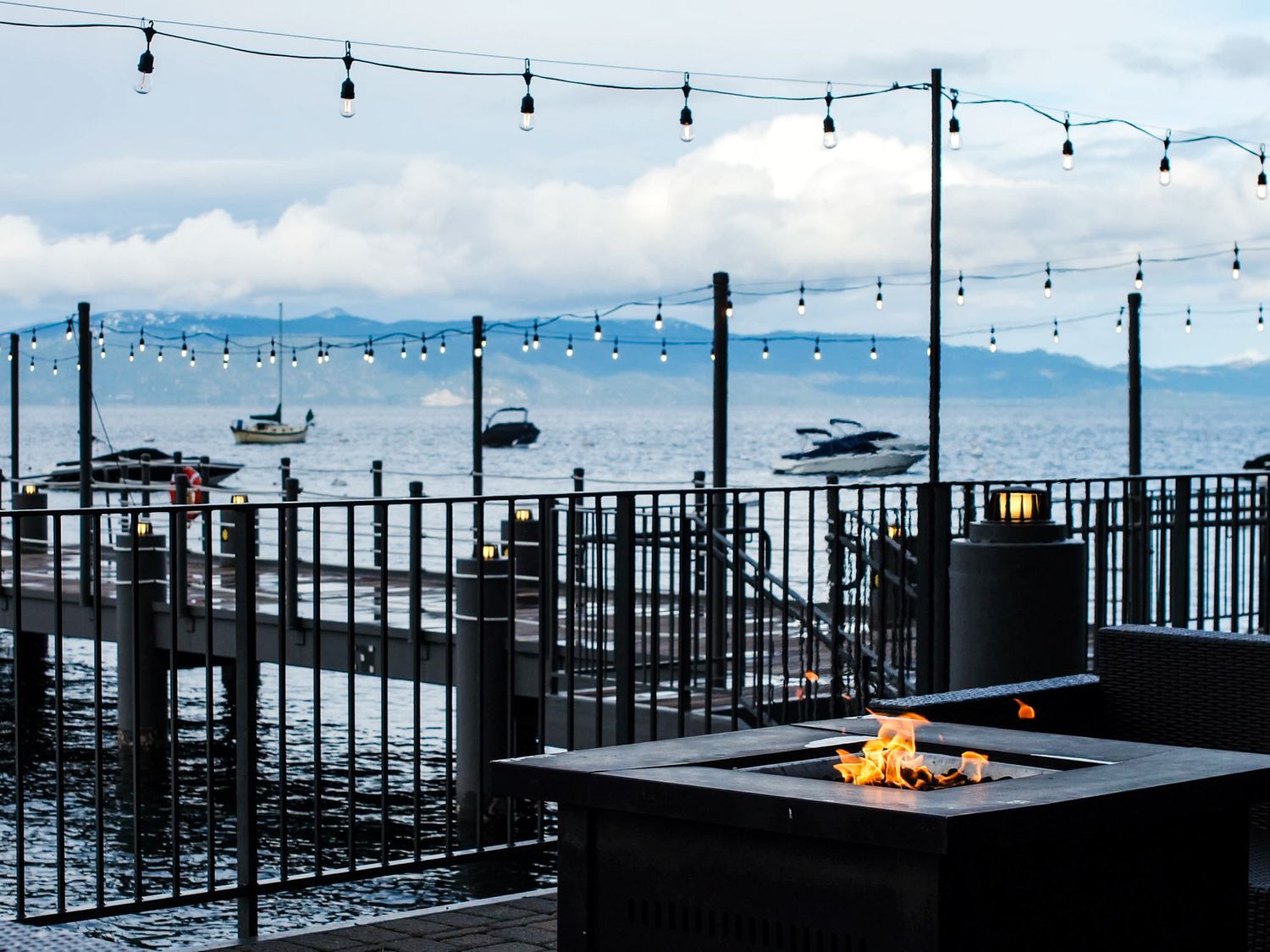 Lakeside patio with string lights and fire pit overlooking boats on calm blue water during daytime.