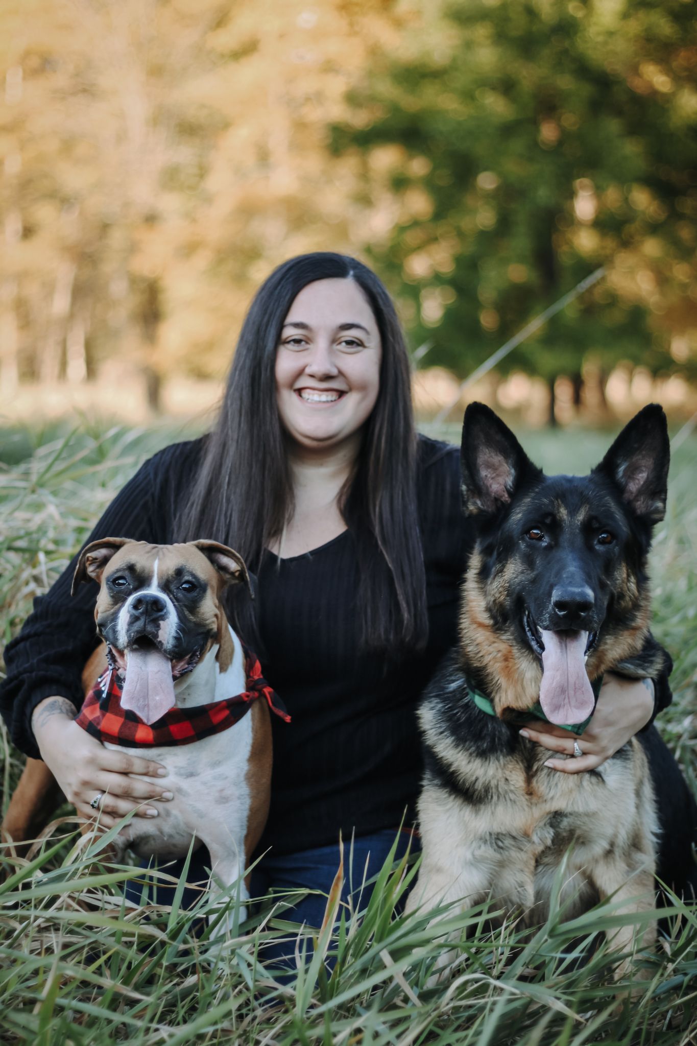A woman and her two dogs posing together in a grassy field at sunset.
