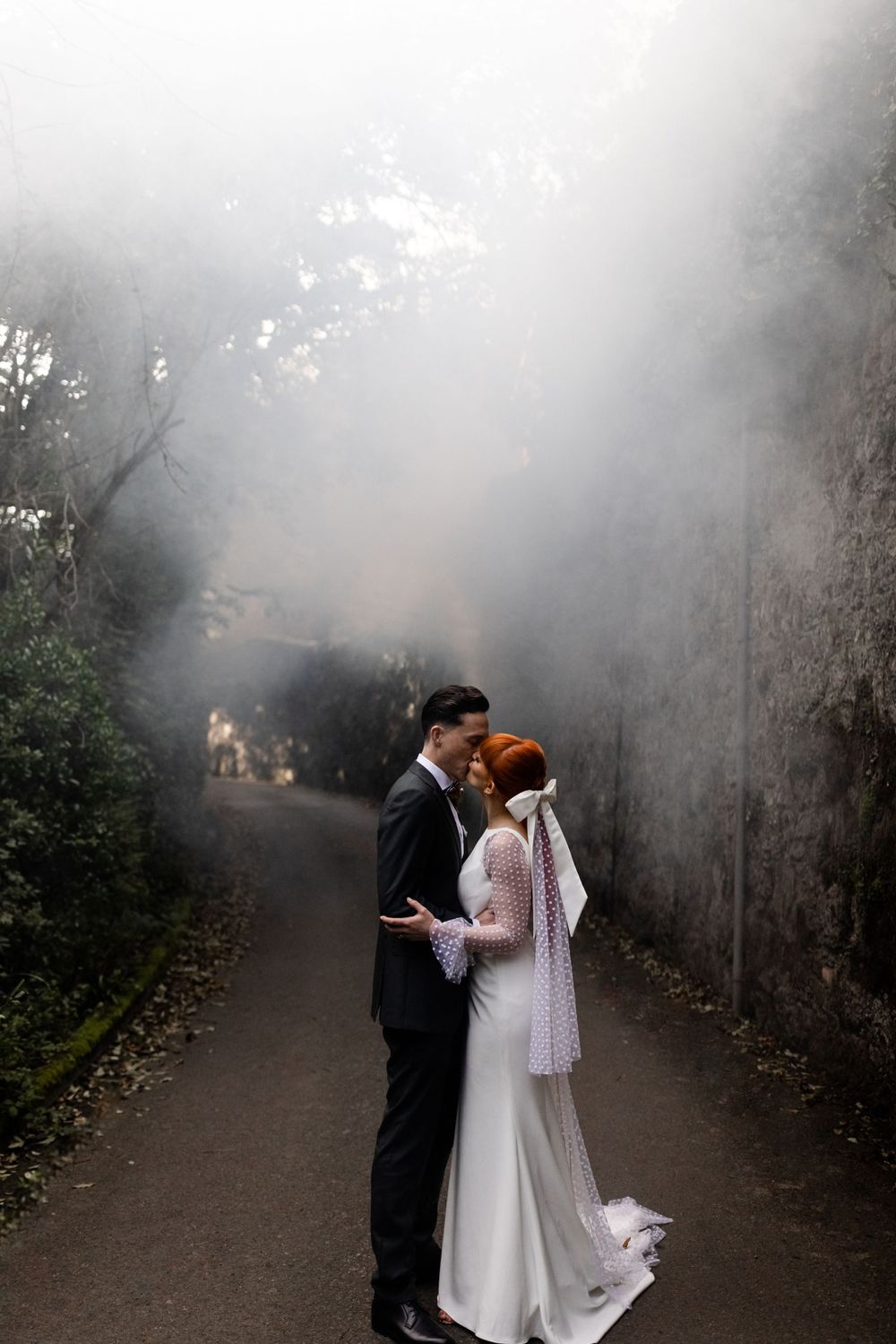 A romantic wedding couple shares a tender moment in a misty garden path surrounded by atmospheric fog.