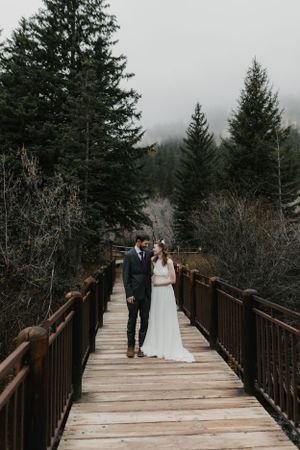 A couple stands together on a wooden bridge walkway surrounded by evergreen trees on a misty winter day.