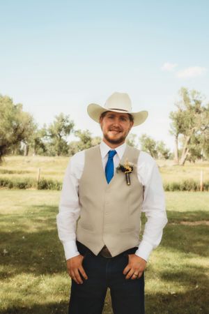 A groomsman in cowboy attire wearing a tan vest and blue tie poses outdoors.