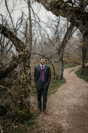 A figure in a dark suit and purple tie stands on a winding dirt path surrounded by bare winter trees in a forest.