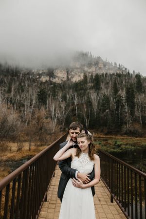 A couple in formal wedding attire embraces on a wooden bridge against a misty mountain backdrop with tall evergreen trees.