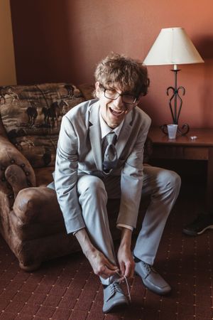 A person in a light gray suit sits on a brown couch, adjusting their shoes.