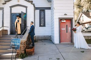 A first look is interrupted by the priest on the steps of a white church building with blue and red doors.