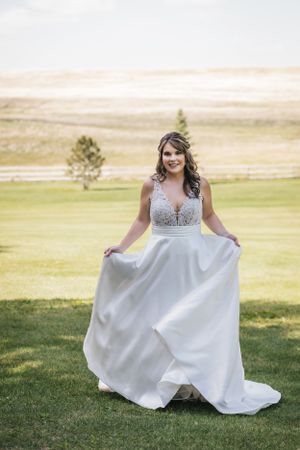 A bride in a flowing white wedding gown with lace bodice twirls on a grassy hillside with scenic views in the background.