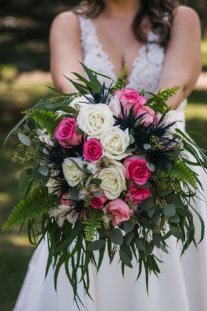 A bridal bouquet with pink and white roses mixed with dark blue thistle and green ferns against a white dress.