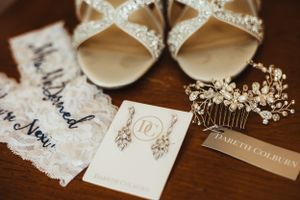 Elegant bridal accessories including sparkly heels, lace garter, crystal hair comb and delicate earrings on wooden surface.
