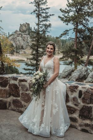 Bride with cascading bouquet poses against mountain backdrop with lake and stone wall.