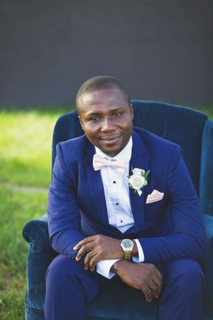 A formal portrait of a groom wearing a navy blue tuxedo and pink bow tie seated in a velvet chair outdoors.