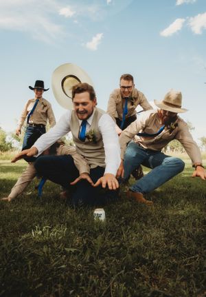 Groomsmen in western attire playfully pose in a field under blue sky.