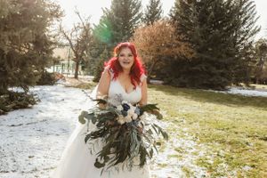 A bride in white holds a unique evergreen bouquet against a snowy winter landscape.