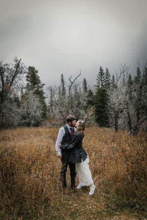 A couple embraces in a moody autumn field surrounded by bare trees and dry grass on an overcast day.