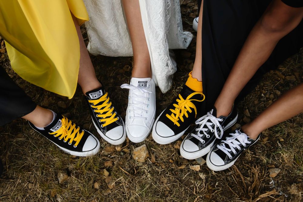 A collection of feet wearing black, white and yellow Converse sneakers positioned in a circle on dirt ground.