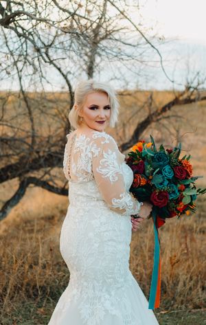 Bride in lace mermaid wedding dress poses with vibrant burgundy and orange bouquet against autumn landscape.