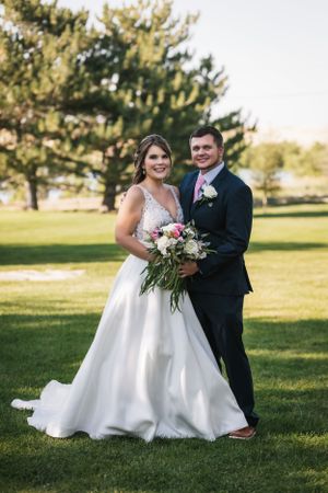 Wedding couple standing on sunny lawn with trees in background as bride holds pink and white floral bouquet.
