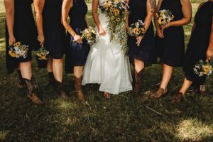 A wedding party showing bridesmaids in navy dresses and brown boots standing with the bride in white on grass.