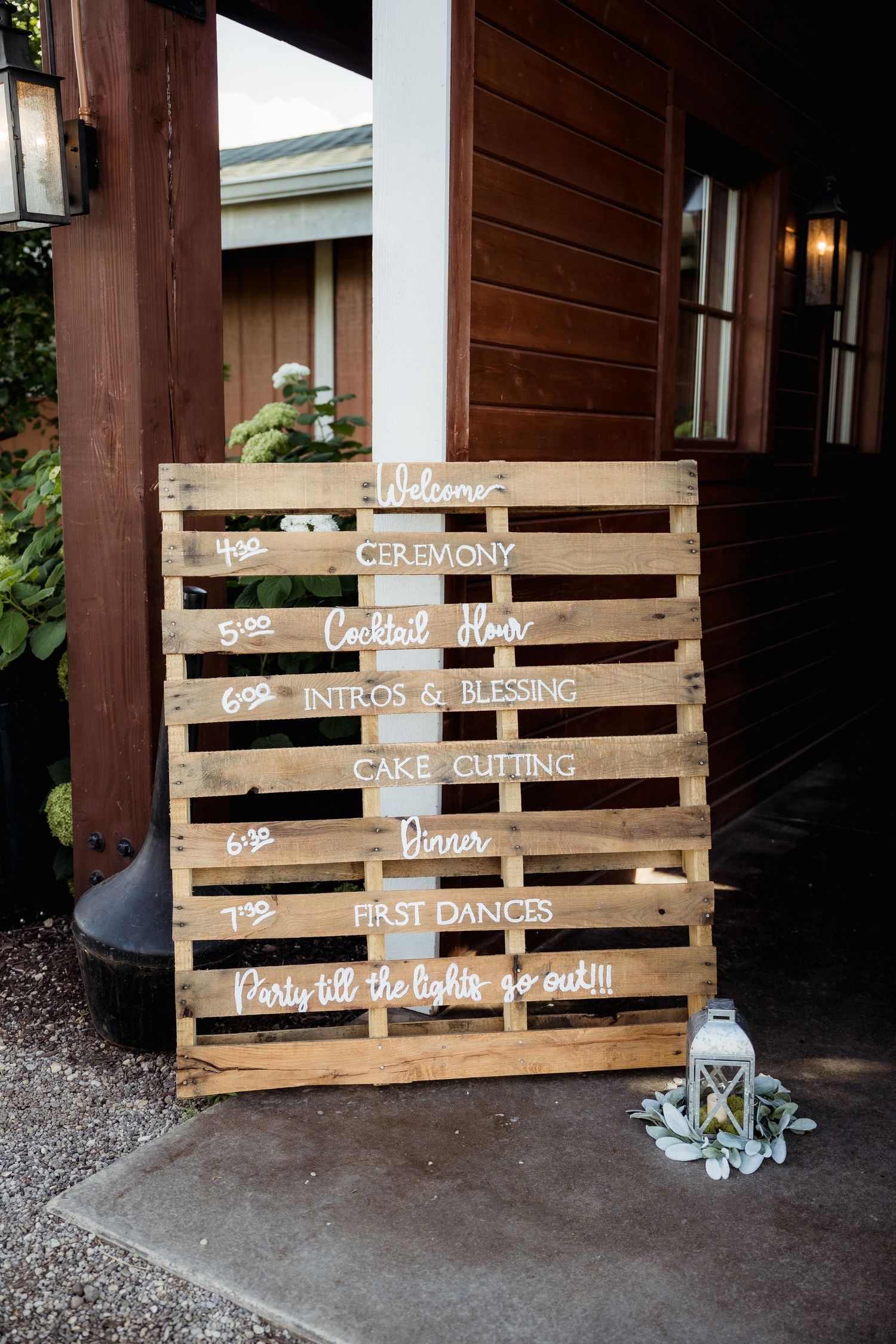 Rustic wooden pallet wedding sign displays event timeline against brown building exterior.