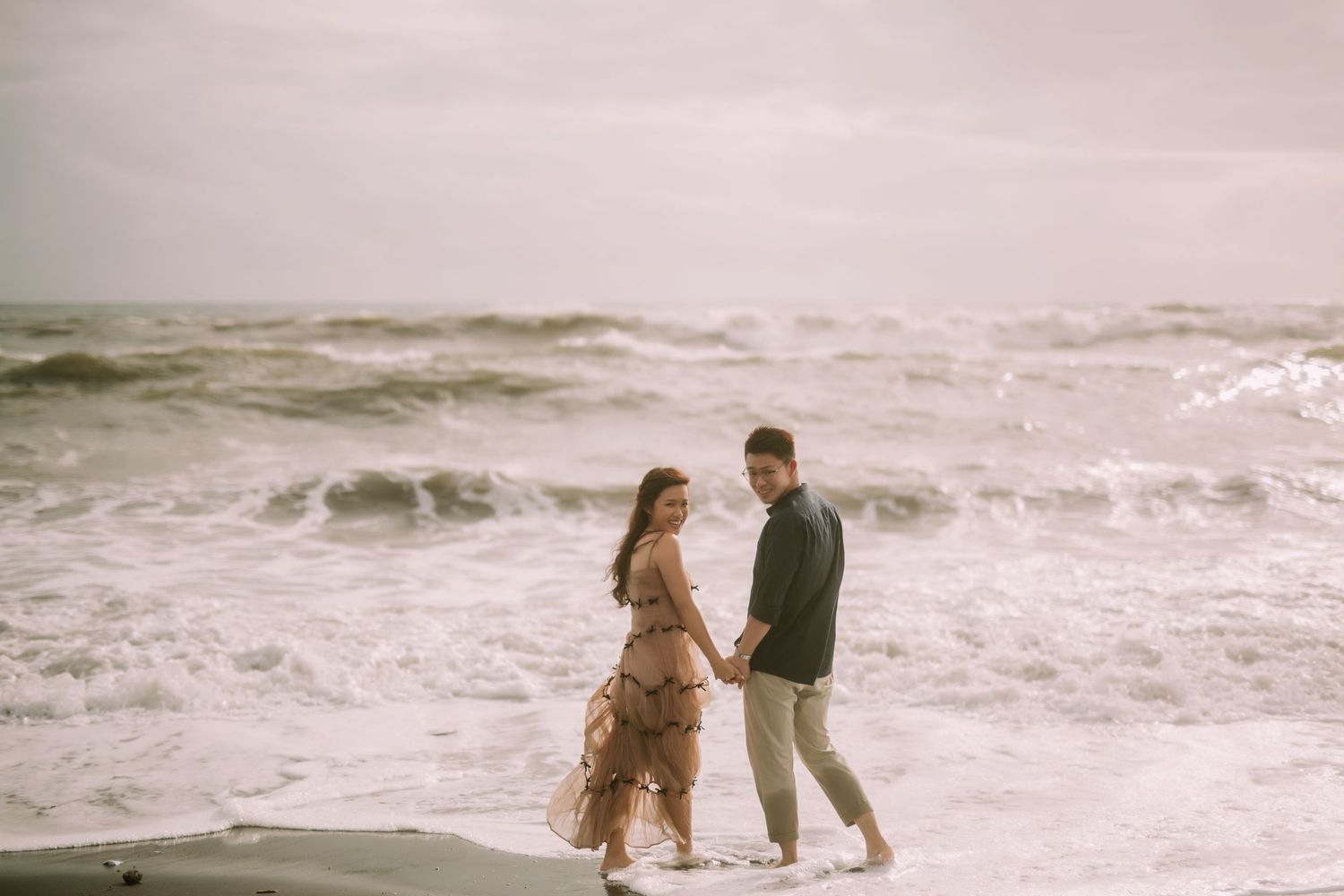 A couple walks along a dramatic coastline with crashing waves and rocky shores at sunset.
