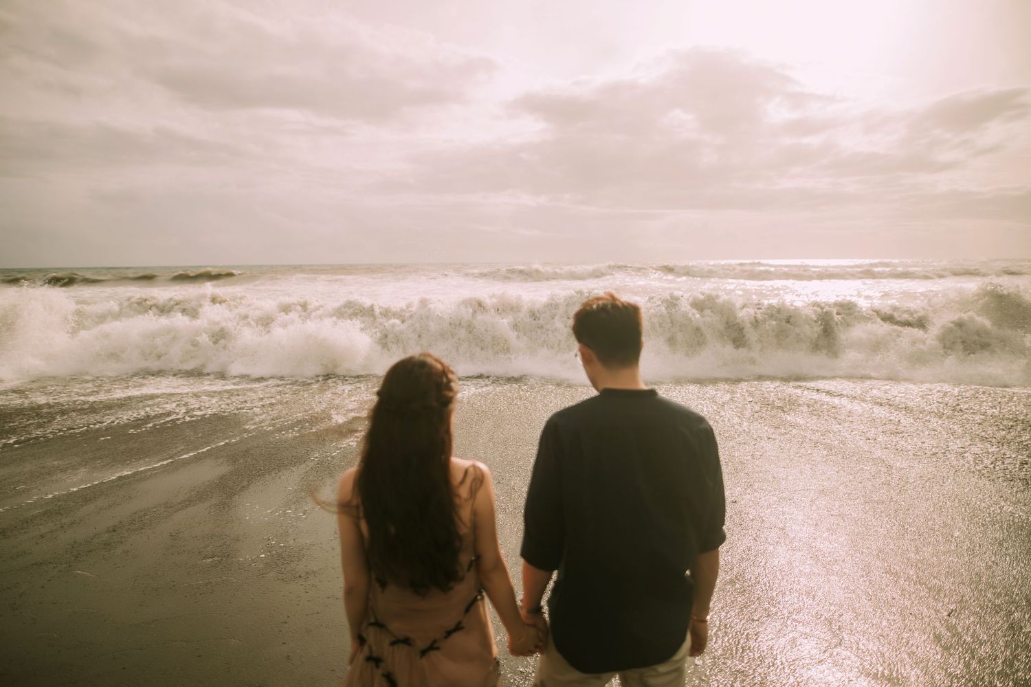 Two people stand together watching waves crash on a moody beach at dusk.