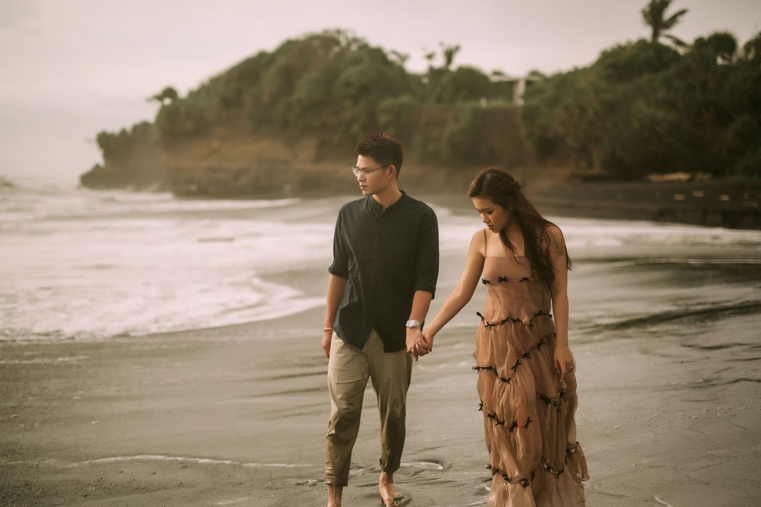 A couple strolls hand in hand along a black sand beach with lush coastal cliffs in the background.