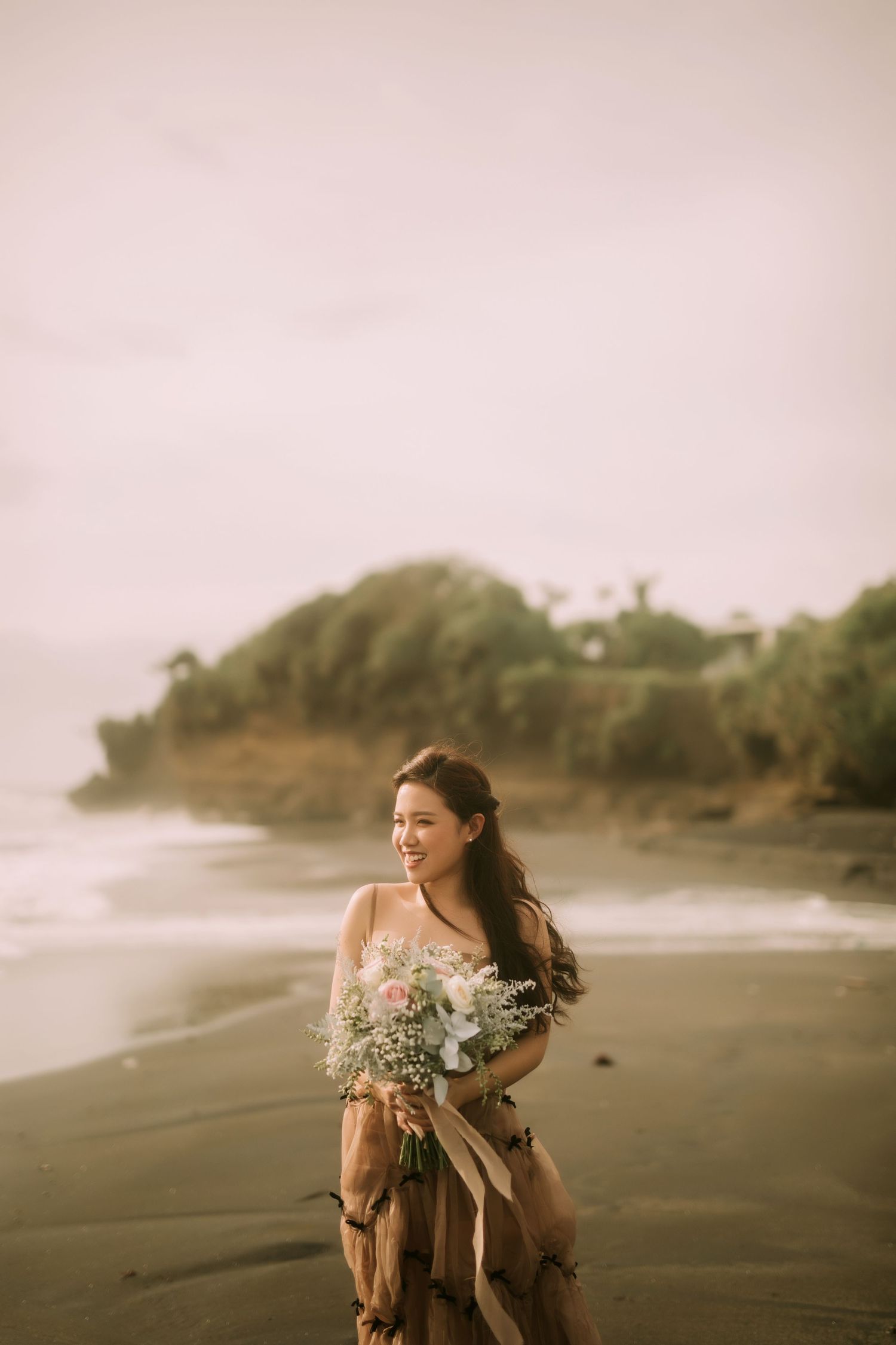 A person in a flowing dress holds a delicate bouquet while standing on a misty beach at sunset.