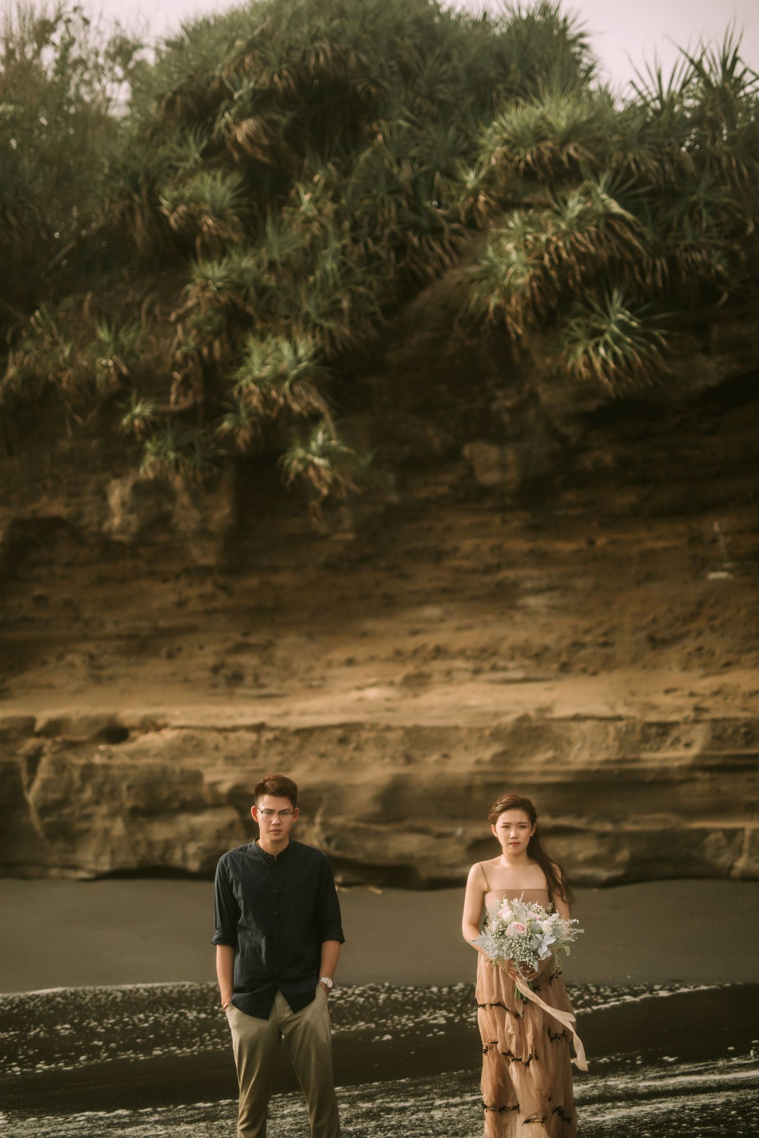 A couple shares an intimate moment on a black sand beach with dramatic cliffs and foliage overhead.