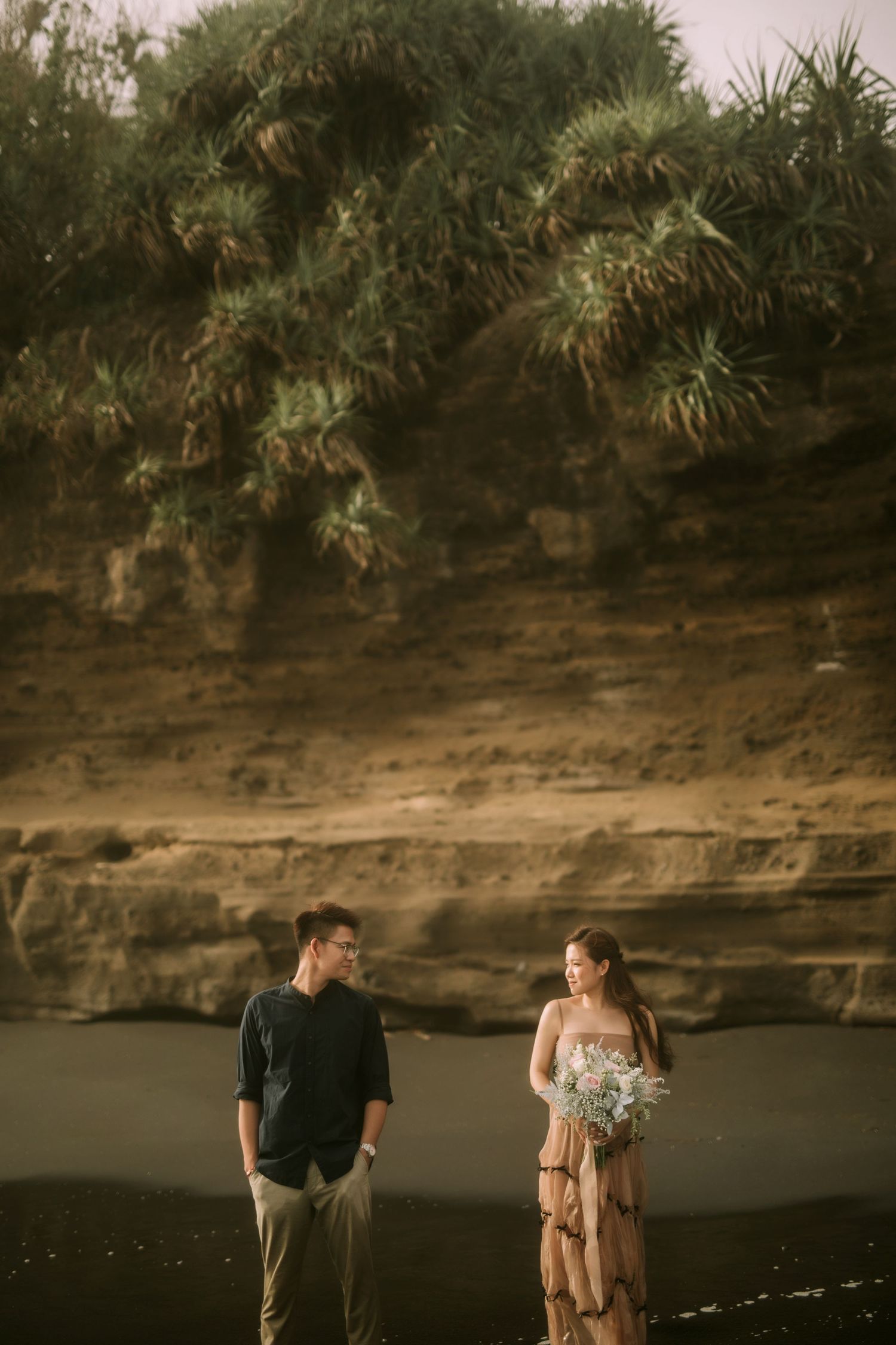 Two people stand beneath towering coastal cliffs on a dramatic black sand beach.