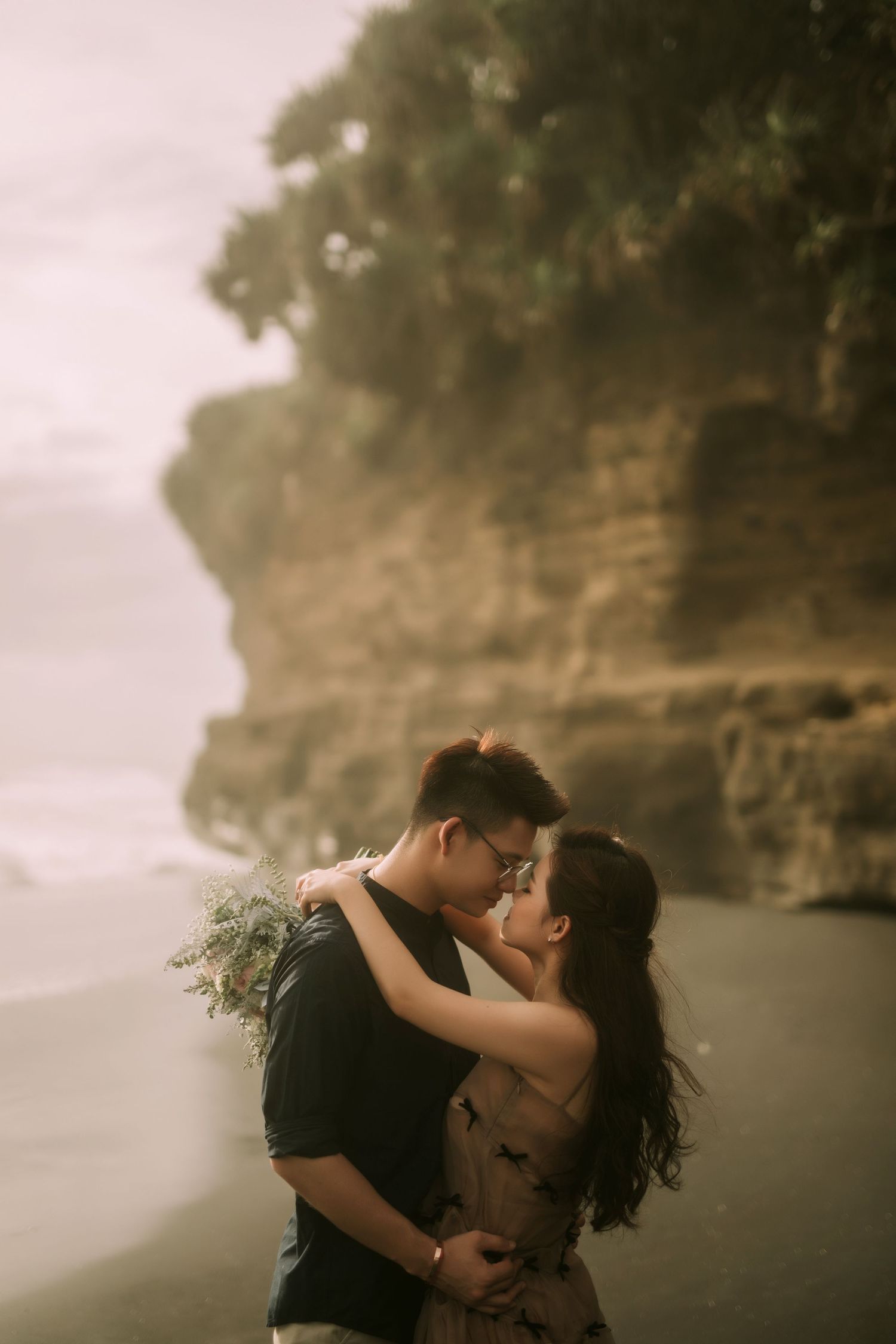 Intimate couple shares a tender moment against moody coastal cliffs and ocean backdrop.