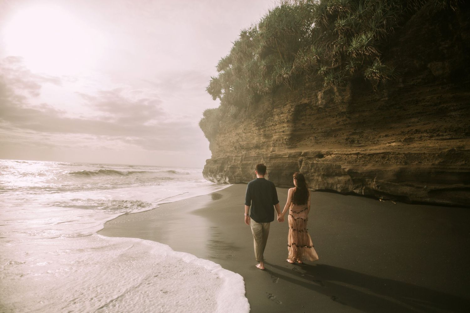 A romantic couple embraces on a misty beach with dramatic cliffs and waves in the background.