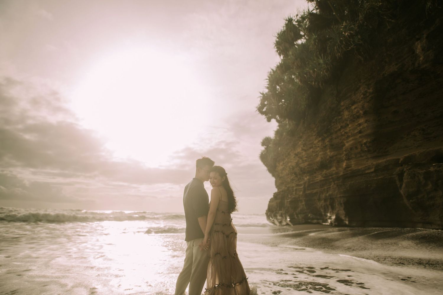 Silhouettes embrace against a misty oceanfront with towering cliffs.