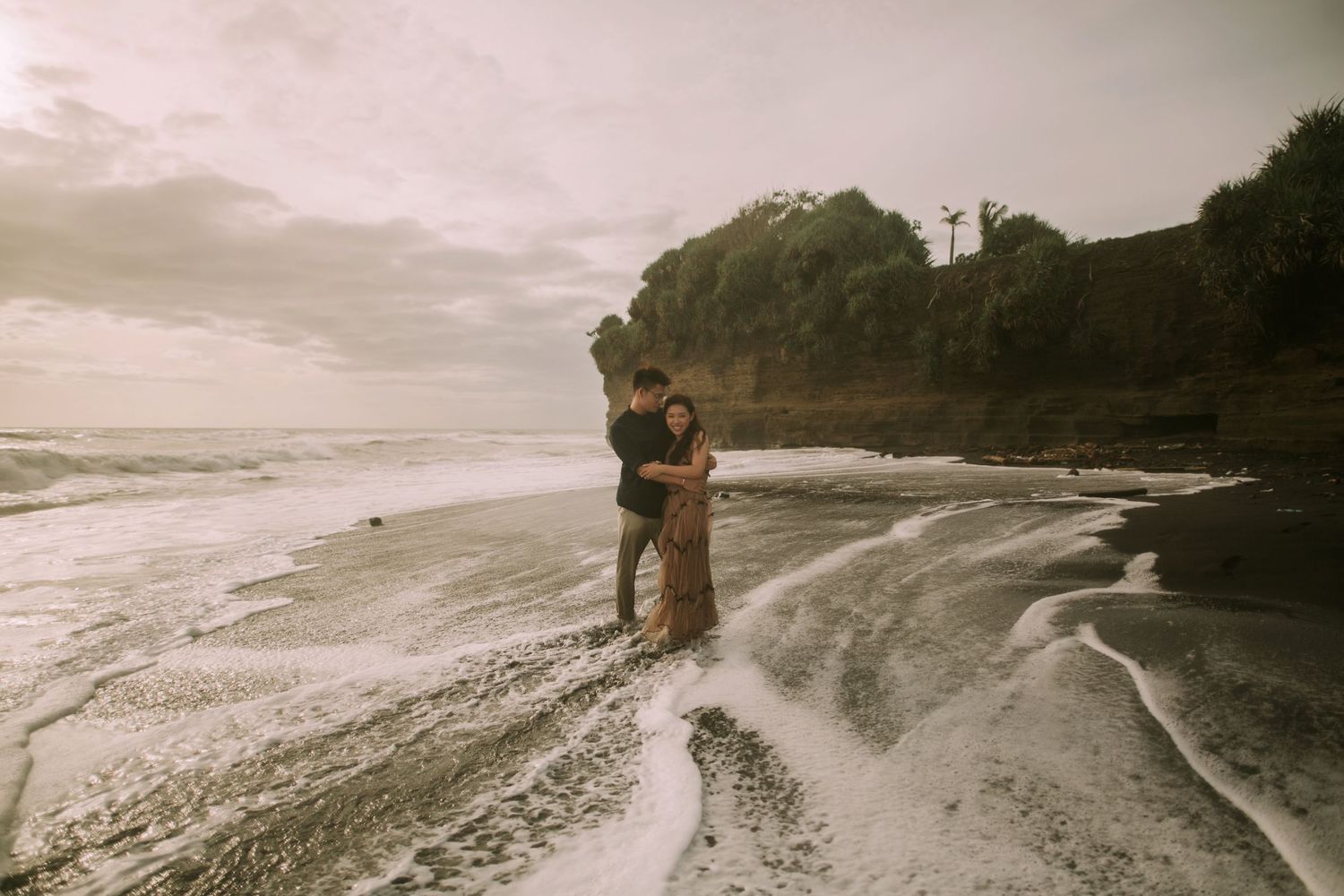 Couple walking along dramatic coastline with waves crashing in sepia tones.