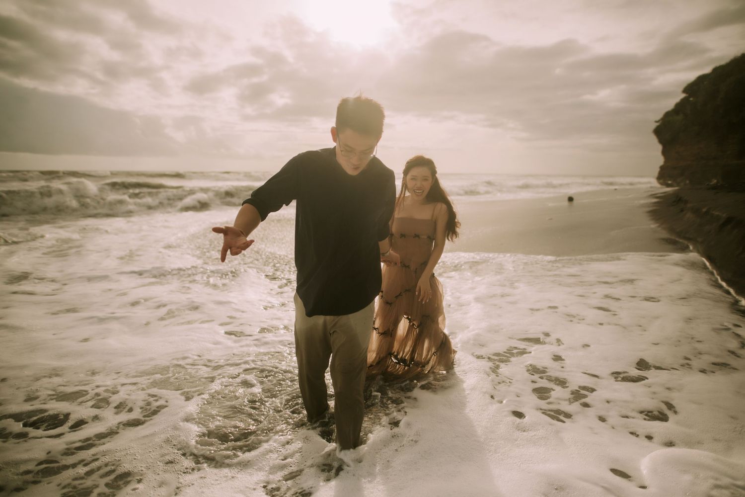 Couple playing in ocean waves along a dramatic rocky coastline.