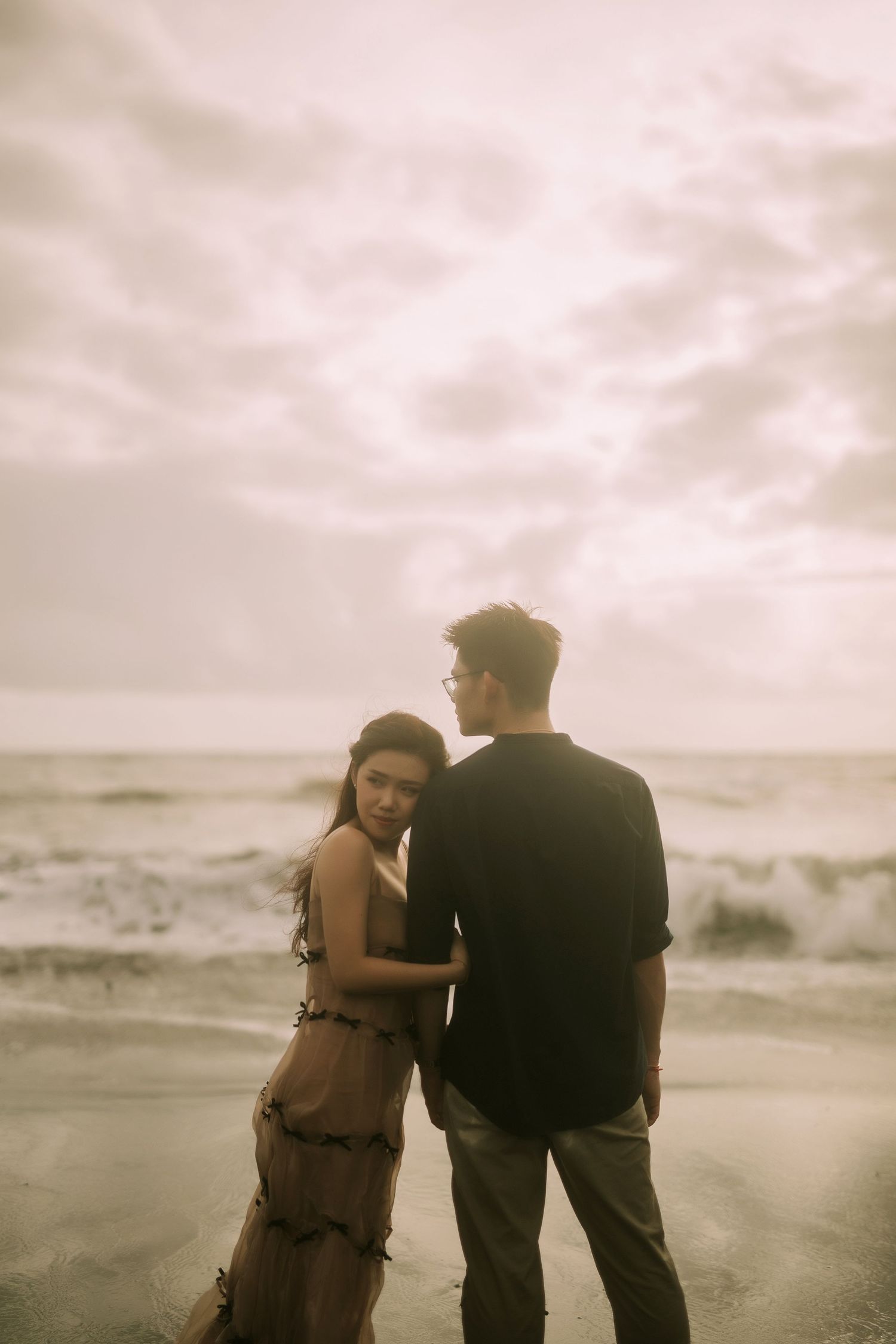 Two figures stand close together on a dramatic beach shoreline at sunset.