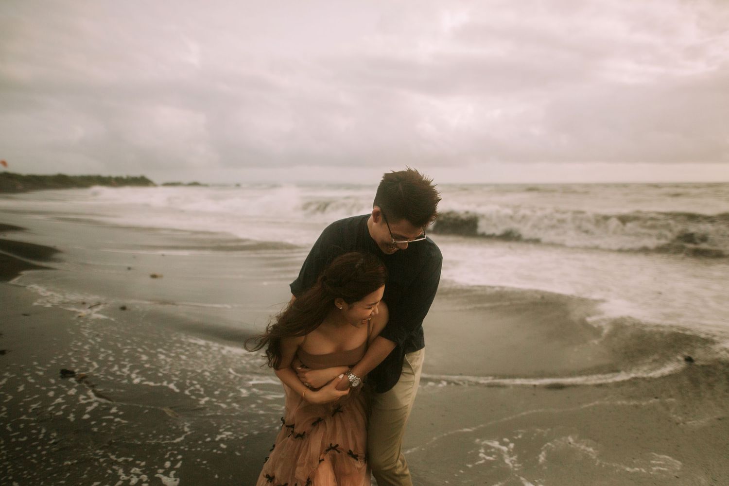 A romantic couple embraces on a moody beach with crashing waves and cloudy skies in the background.