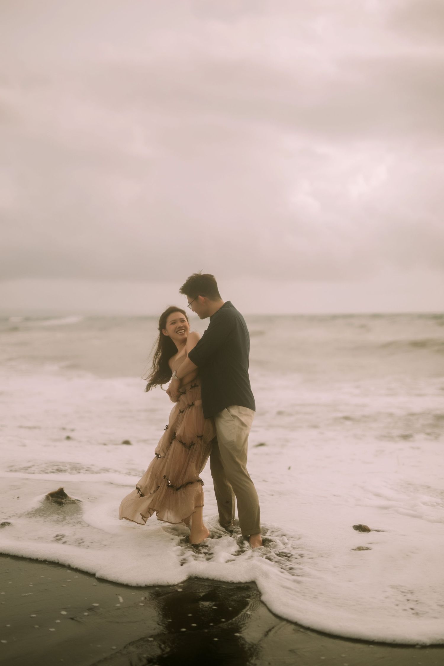 A couple shares an intimate dance-like moment on the beach as waves crash behind them.