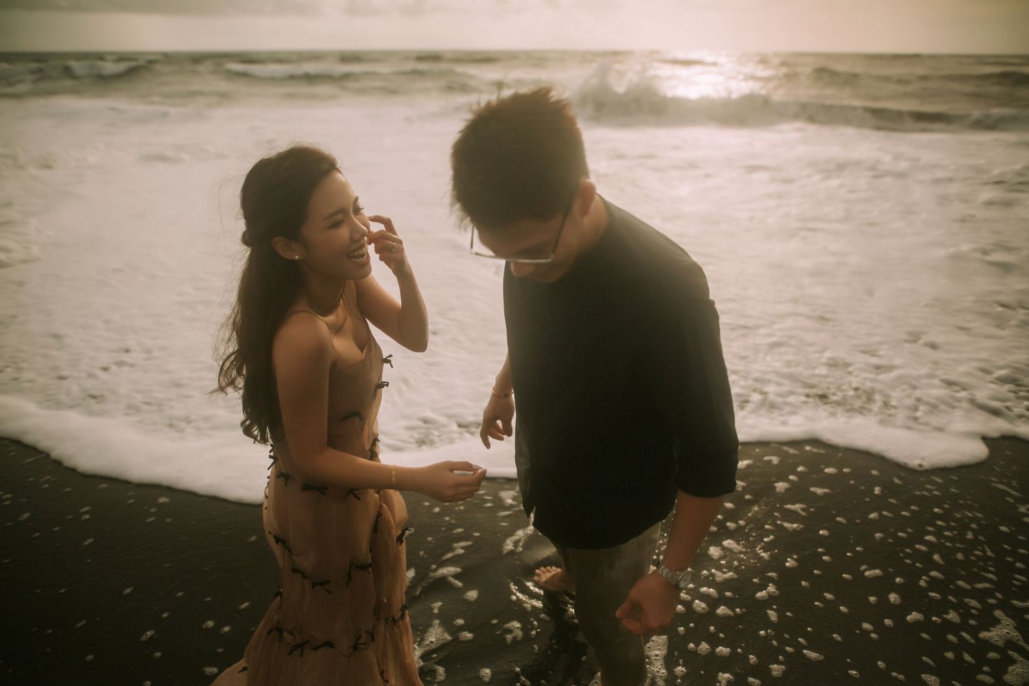A couple interacts joyfully on a dark sandy beach with waves in the background.