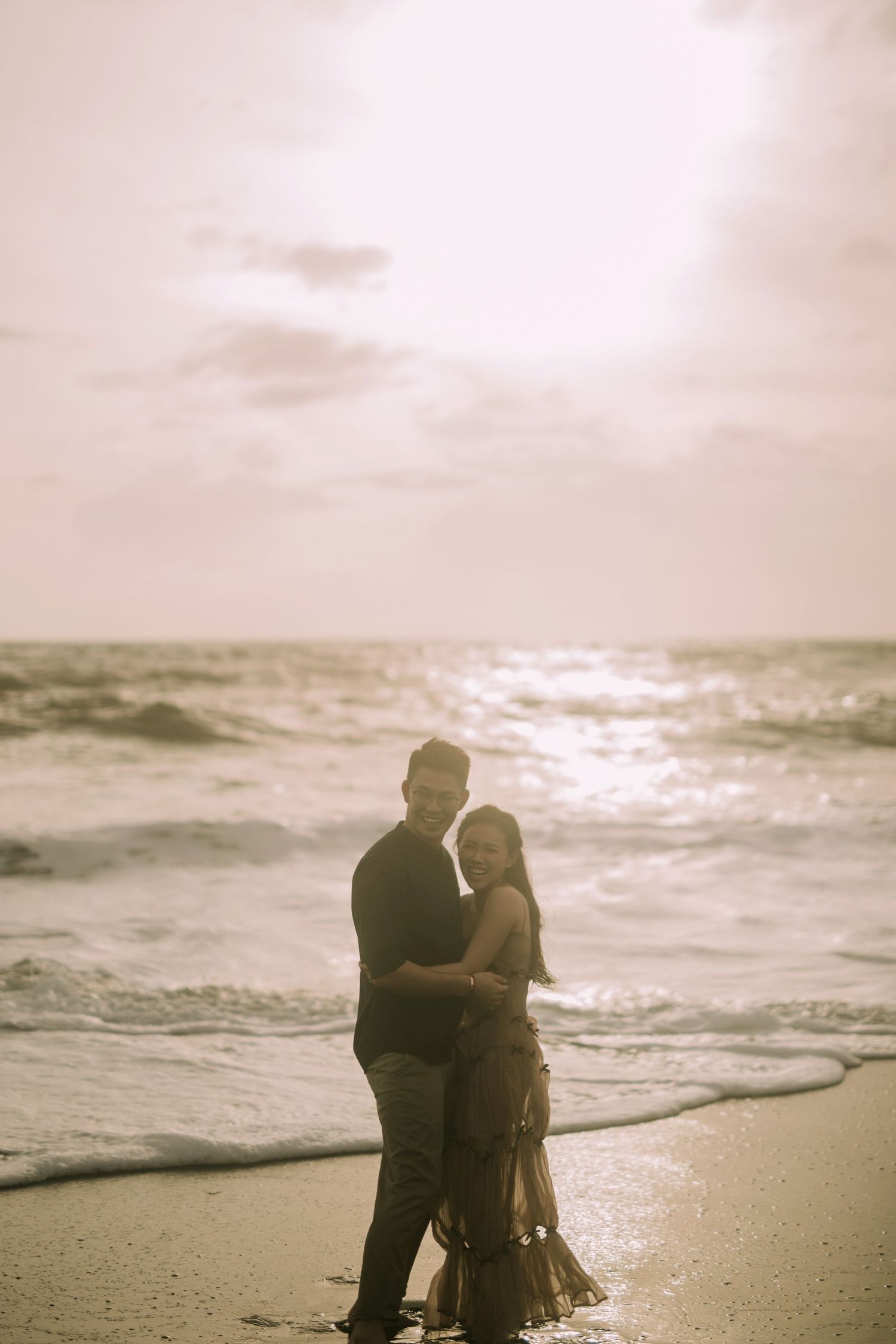 A couple shares an intimate moment on the beach at sunset with waves crashing behind them.
