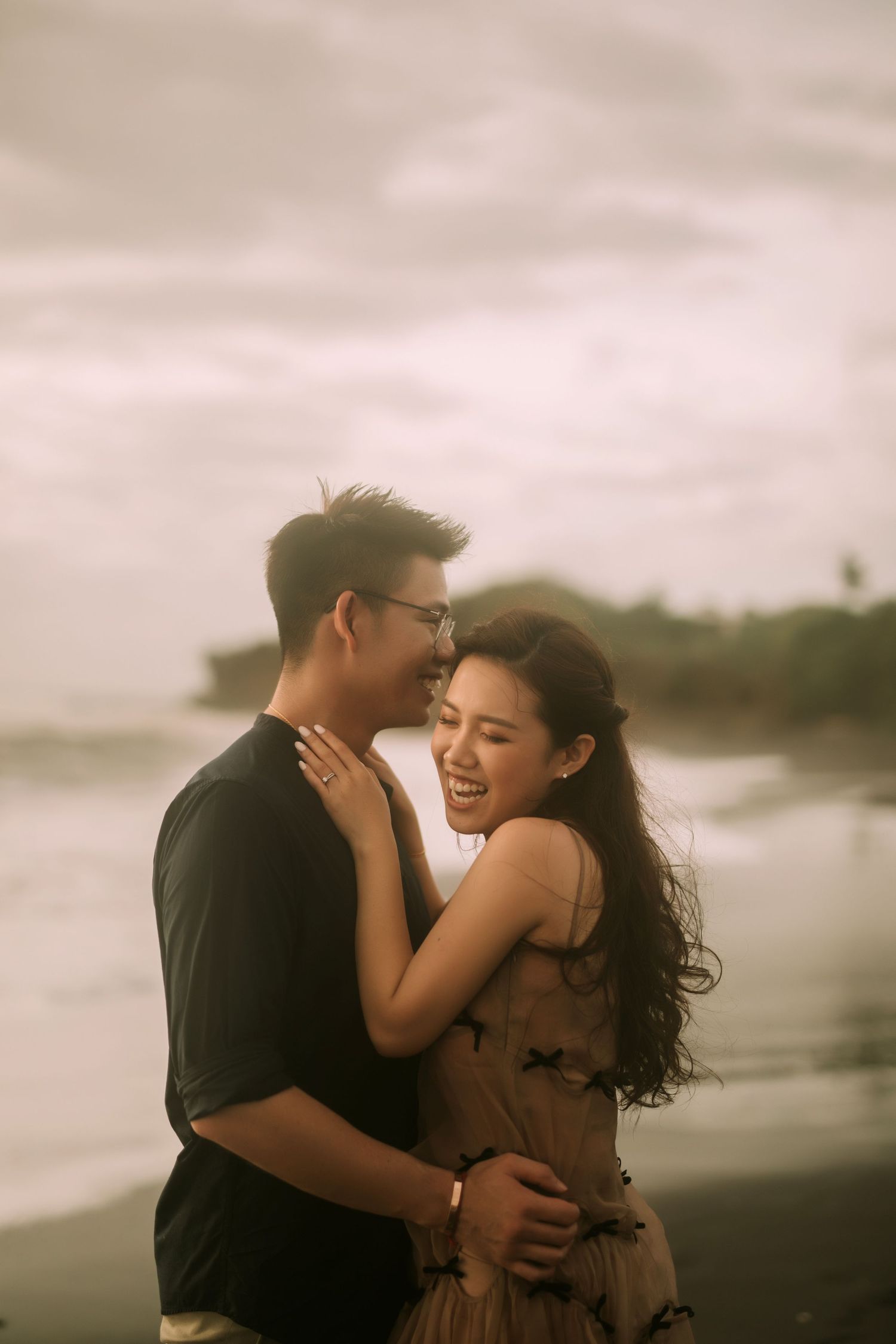 A romantic couple embraces and laughs together on a moody beach at sunset.