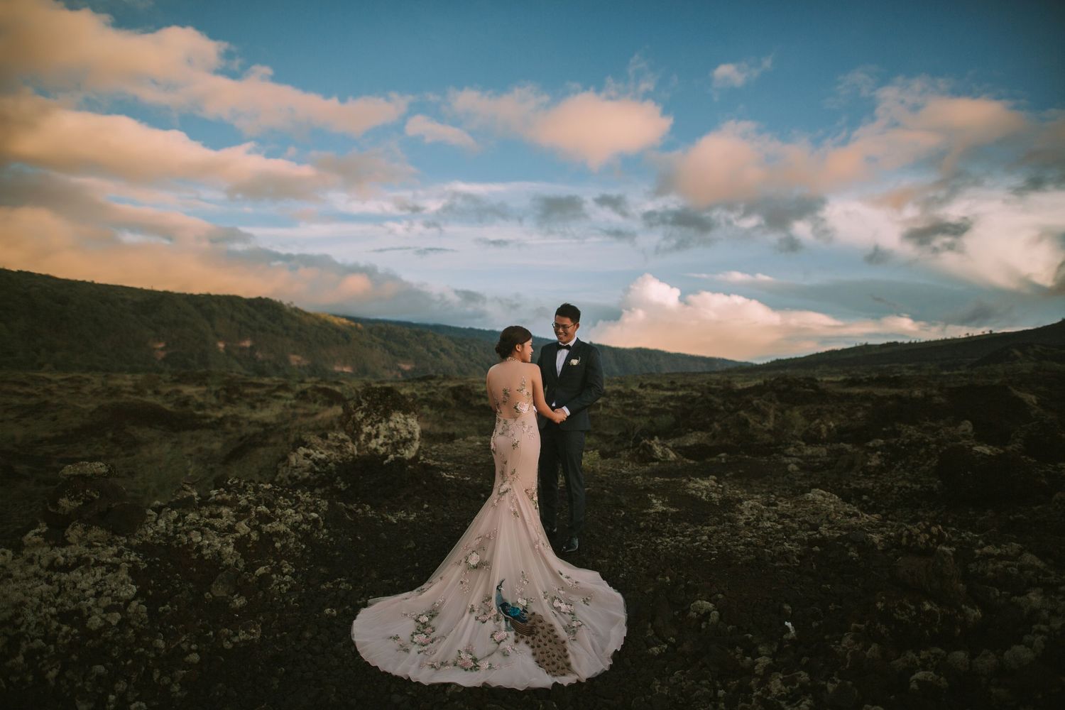 Bride in flowing dress stands with partner against mountainous landscape.