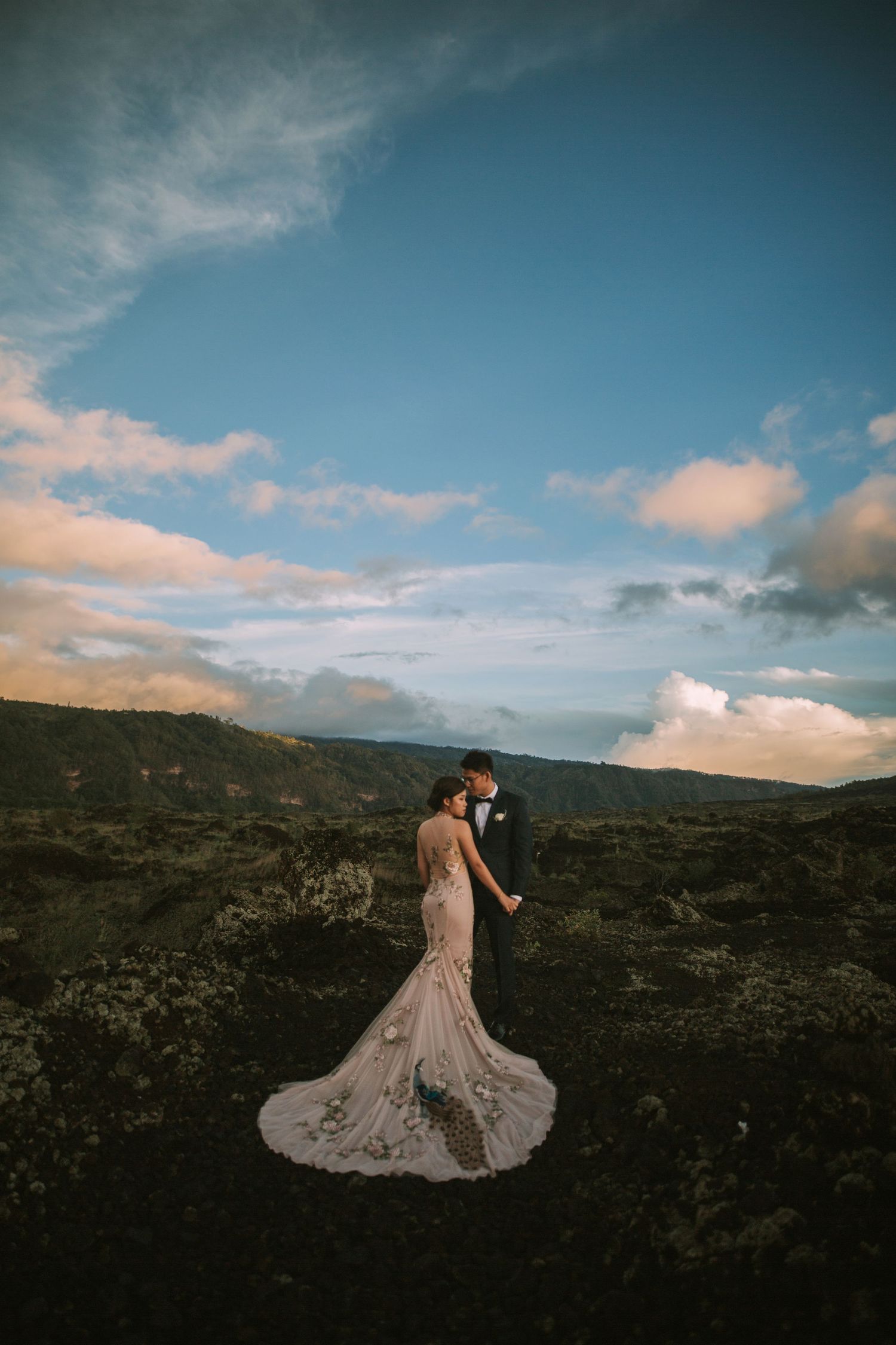 Elegant wedding couple embraces on rocky terrain with dramatic sky backdrop.