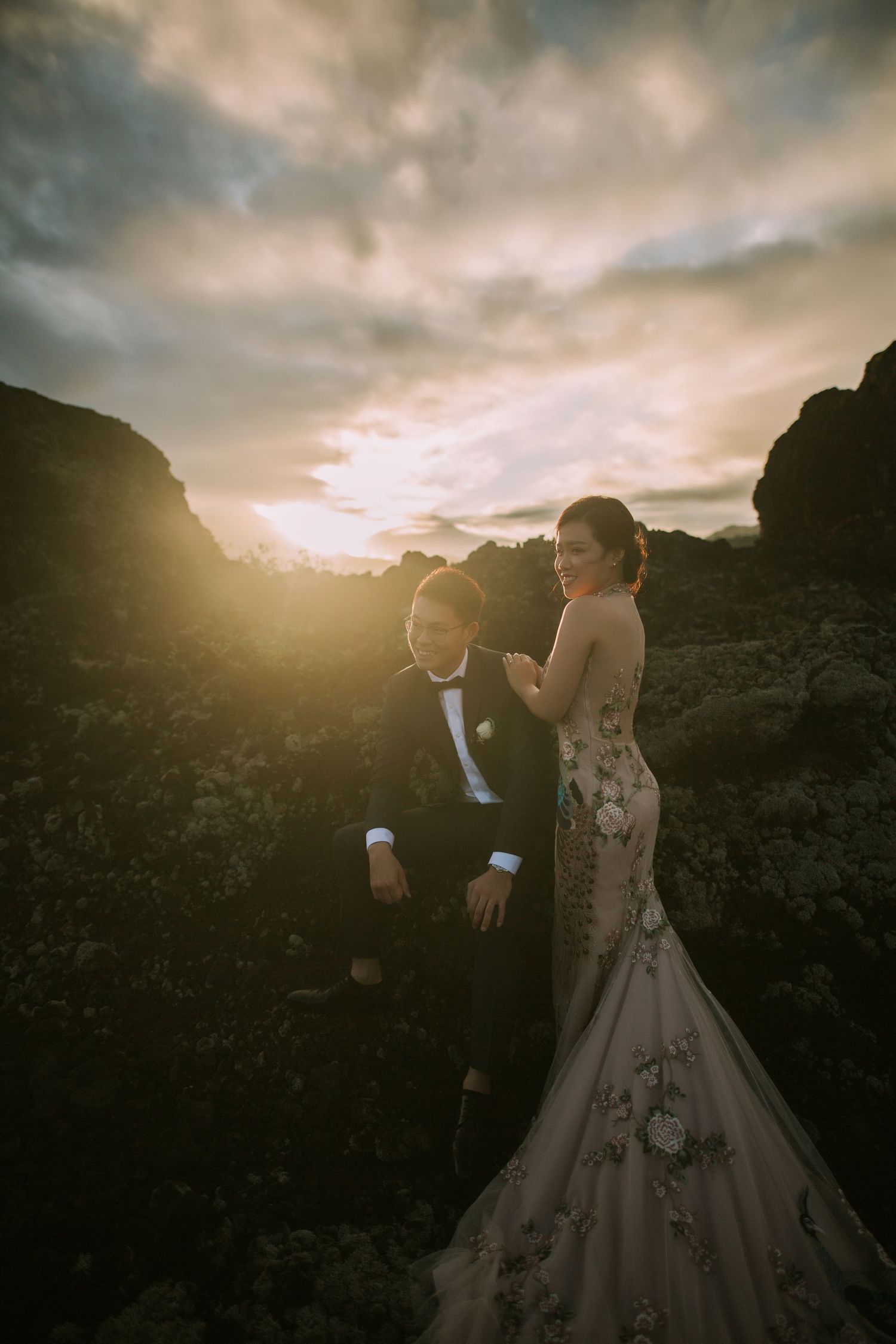 Couple in formal attire walks together against dramatic sunset landscape.