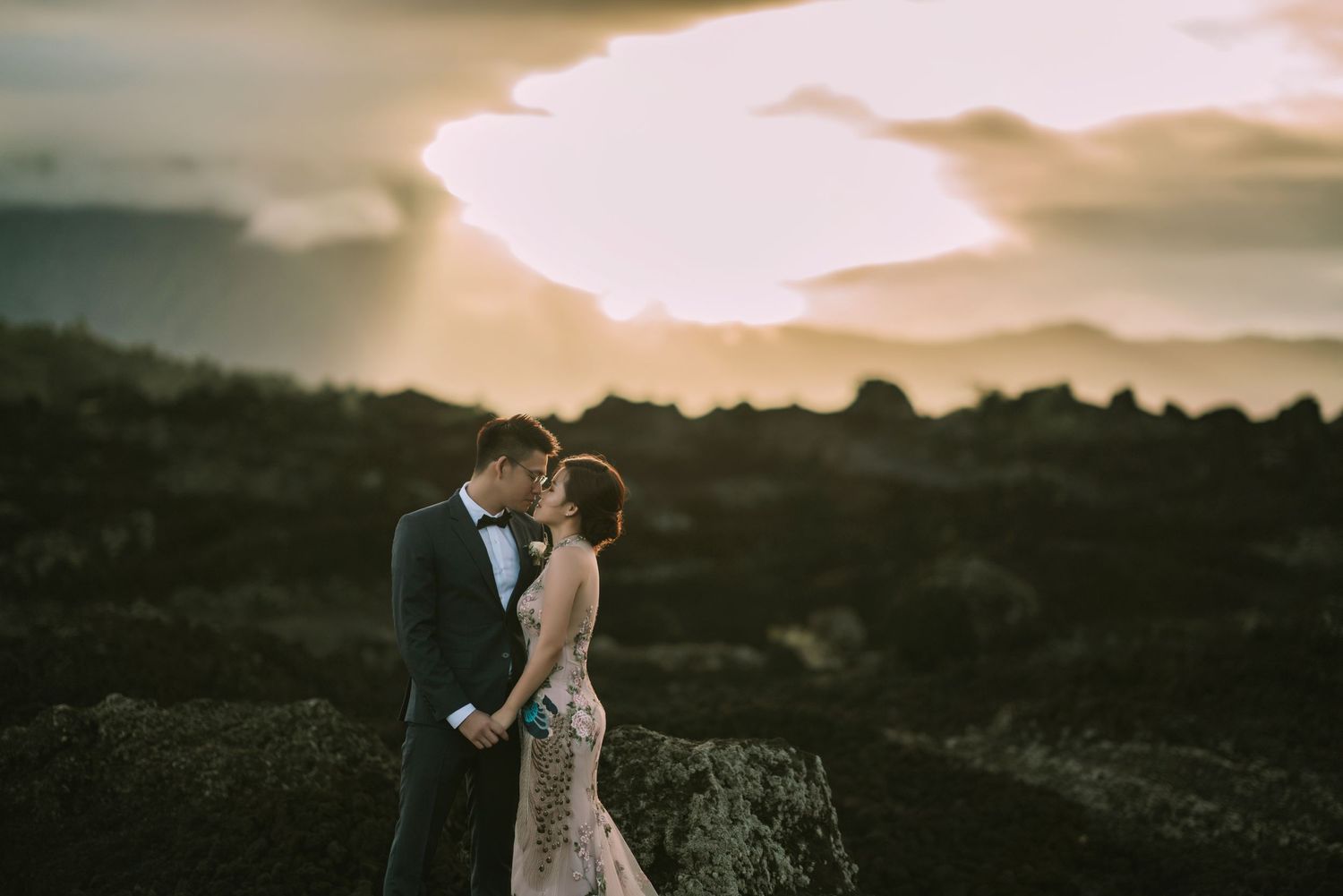 Couple in wedding attire embrace against a dramatic mountain sunset backdrop.
