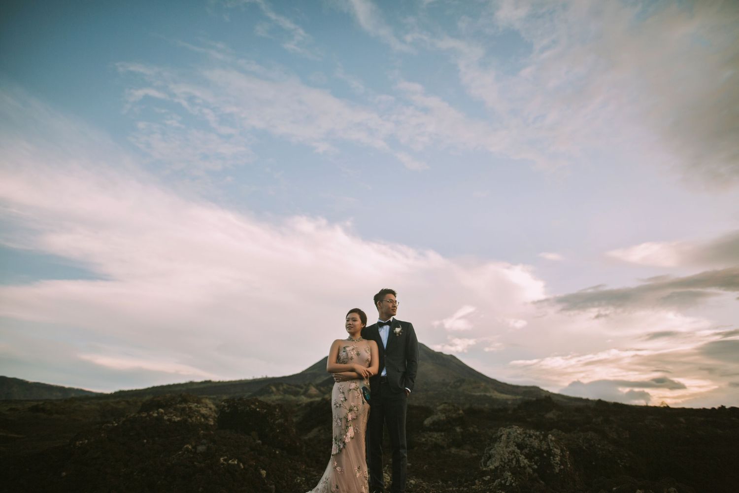 Wedding couple stands together against mountain landscape and cloudy sky.