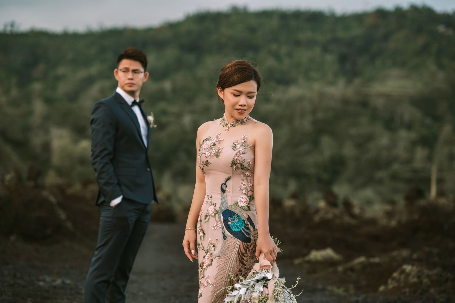 Couple poses for romantic wedding photos on rocky volcanic terrain with lush green mountains in background.