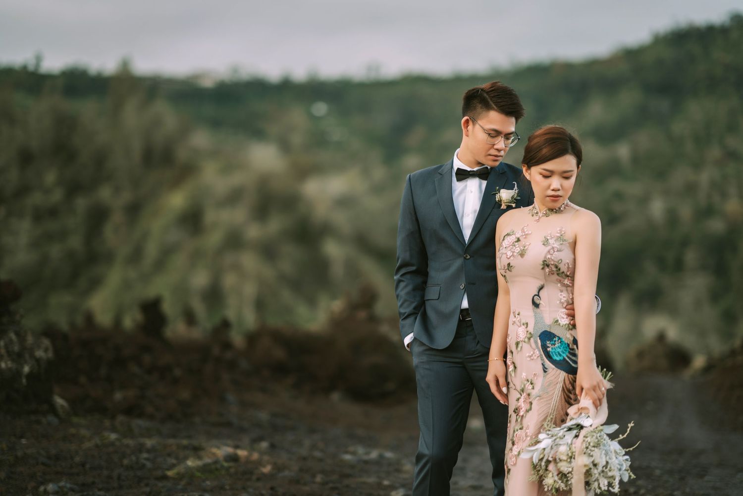 Wedding couple stands together on dark volcanic rocks with misty mountains behind them.